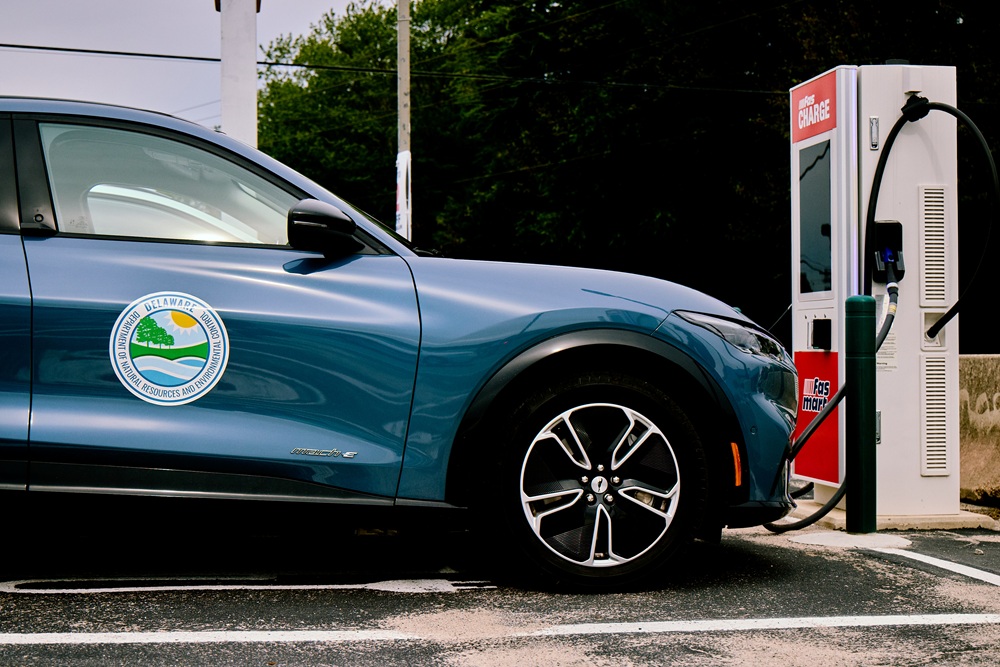 A blue car with the Delaware DNREC logo sits at an electric charging station.